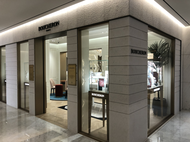 Eintritt in ein Boutique in einem Einkaufszentrum mit Glaswänden und -türen, Text an den Wänden, Deckenleuchten, Stühlen, Tischen, einer Pflanze und anderen Gegenständen, mit sichtbarem Boden.