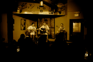 Eine Gruppe von Musikern in weißen Hemden, die Gitarren spielen und in ein Mikrofon singen, auf einer Bühne mit einem Schlagzeug dahinter, Gemälden an den Wänden, einer Tür mit einem Schild auf der rechten Seite, einem Banner über der Bühne und einem Publikum davor.