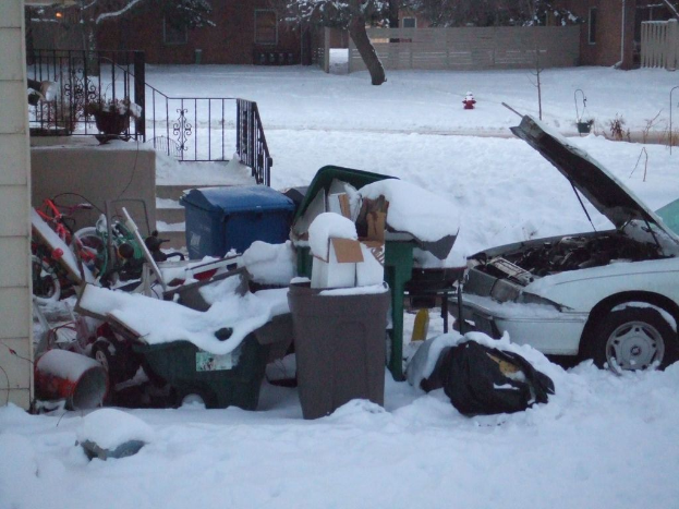Eine Außenaufnahme mit Müll in der Mitte, einem Auto auf der rechten Seite, Treppen auf der linken Seite, Bäumen oben und Eis am Boden.