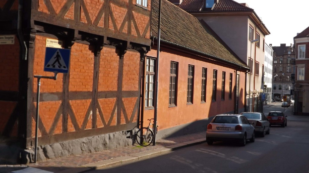 Fahrzeuge auf der Straße mit Gebäuden, Laternen, Verkehrsschildern, Fenstern und Dächern im Hintergrund und einem Fahrrad auf dem Gehweg in der Nähe eines Pfahls.