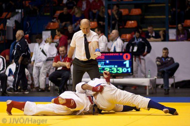 Zwei Judokämpfer im Match mit einem Schiedsrichter und Zuschauern in der Nähe von Geländern, Treppen und einer Anzeigetafel.