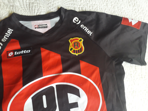 Schwarze und rote Fußballtrikot mit einer roten und schwarzen Streifen, mit einem Logo und Text, auf einer weißen Oberfläche angezeigt.
