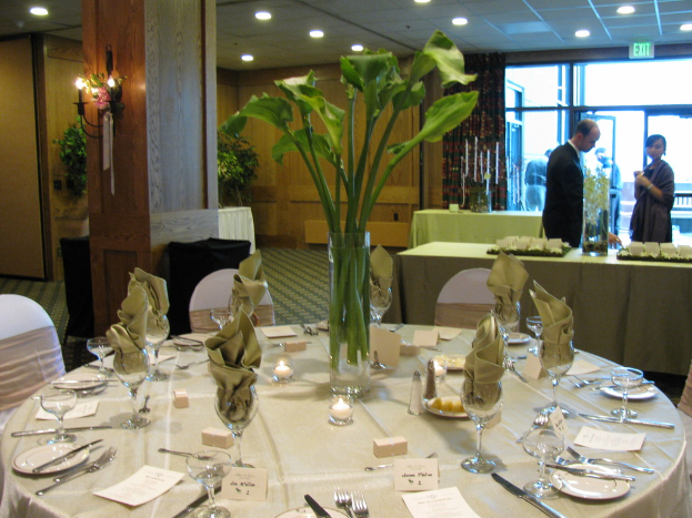 Ein Tisch mit einer weißen Decke, Tellern, Besteck und einem Glas, umgeben von Stühlen, mit einem grünen Tisch dahinter, einem Mann in einem schwarzen Blazer, einem stehenden Mädchen, einer Ausgangstür und zwei Männern hinter der Tür.