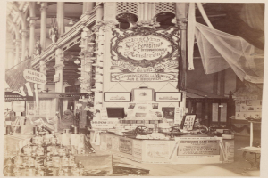 Ein altes Schwarz-Weiß-Foto eines Pariser Ladens mit Flaschen links, Fotorahmen an der Wand und einer Tafel mit Schrift, in einer Ausstellungshalle mit Säulen und einem Netz im Hintergrund.