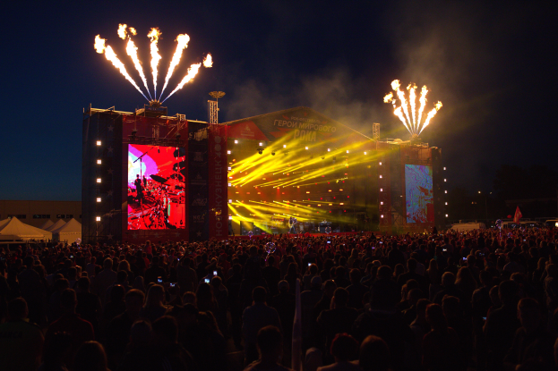 Eine große Menschenmenge auf einem Musikfestival mit einer Bühne in der Mitte, Musiker unter hellen Scheinwerfern und Bannern, Zelte links und ein dunkler Himmel mit Feuerwerk im Hintergrund.