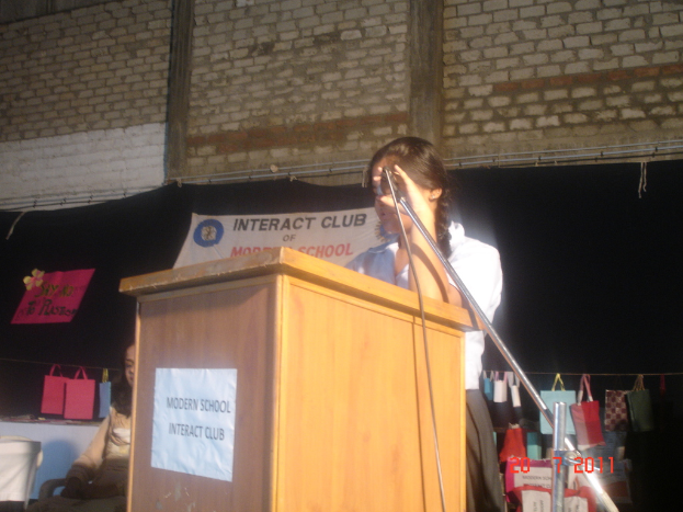 Ein Mädchen in einem weißen Hemd steht neben einem Podium in einem Konferenzraum, mit einem Mikrofonständer rechts daneben, einem weißen Banner an einer Wand im Hintergrund und mehreren Taschen, die an einem Faden rechts hängen.