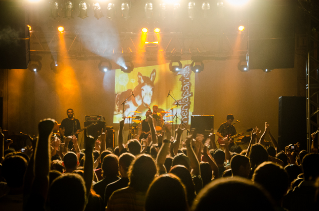 Eine Gruppe von Menschen mit einigen, die Musikinstrumente vor Mikrofonen spielen, umgeben von Lautsprechern, Lichtern und Metallstangen.