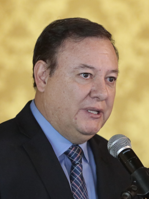 Ein Mann in Anzug und Krawatte, der in ein Mikrofon spricht, identifiziert als Generalstaatsanwalt, mit unscharfem Hintergrund.