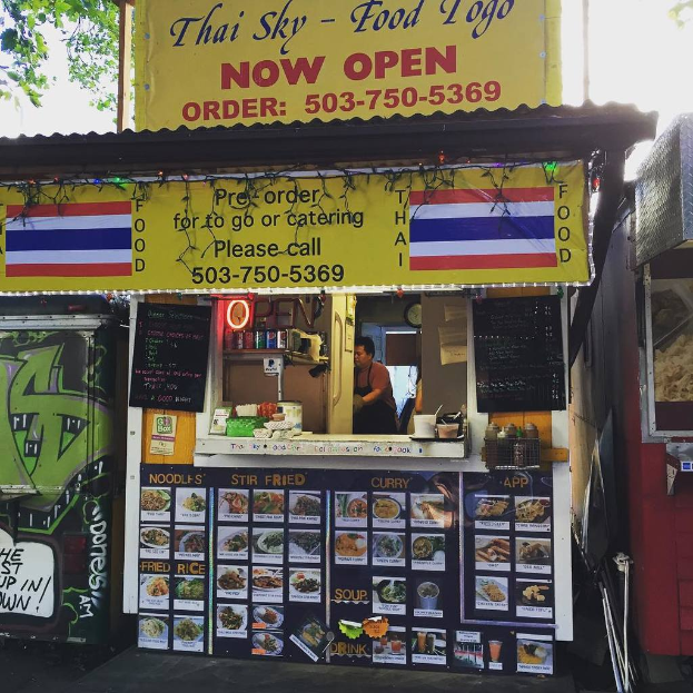 Ein Ladengeschäft mit verschiedenen Artikeln, darunter Getränkedosen, Speisekarten, eine Uhr, Gewürze in Plastikbehältern, Gewürze, Werbetafeln und Namensschilder, vor einem Himmel als Hintergrund.