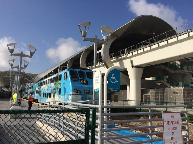 Einige Personen auf einem linken Bahnsteig mit einem Zug auf der rechten Seite, eine Brücke, ein Zaun und mehrere Pfosten.
