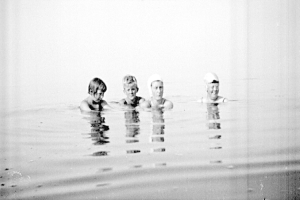 Schwarzes und weißes Foto von vier Kindern, die in einem Pool schwimmen und alle eine Schwimmkappe tragen.