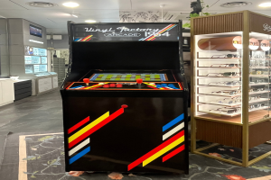 Ein Geschäft mit einem zentralen Retro-Arcade-Automaten, einem Regal mit Gläsern auf der rechten Seite und Hintergrundelementen wie Schränken, einem Fernseher und anderen Gegenständen, mit Deckenleuchten, die eine Retro-Atmosphäre schaffen.