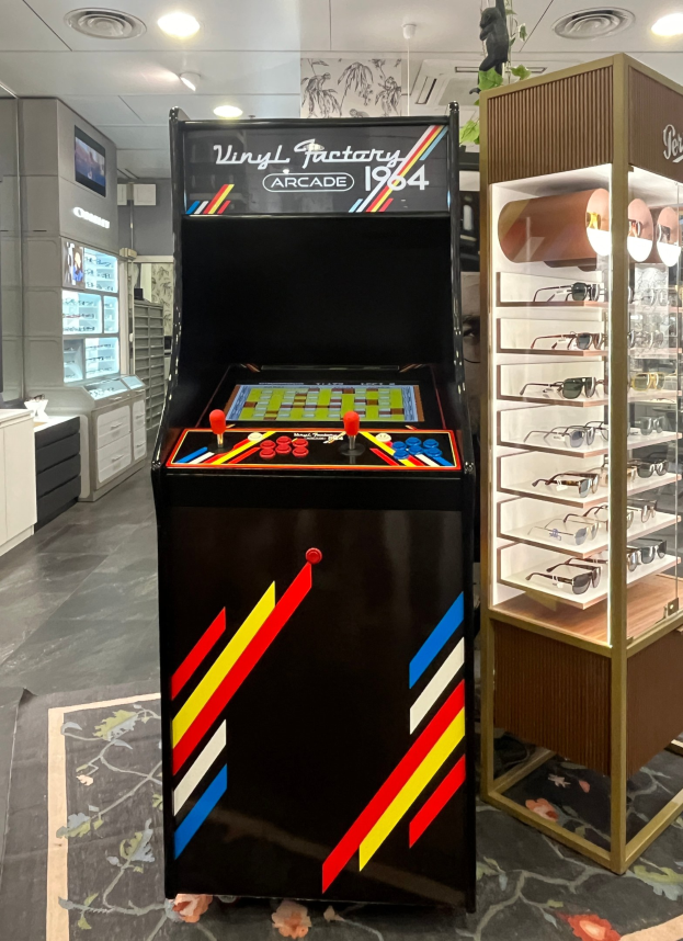 Ein Geschäft mit einem zentralen Retro-Arcade-Automaten, einem Regal mit Gläsern auf der rechten Seite und Hintergrundelementen wie Schränken, einem Fernseher und anderen Gegenständen, mit Deckenleuchten, die eine Retro-Atmosphäre schaffen.