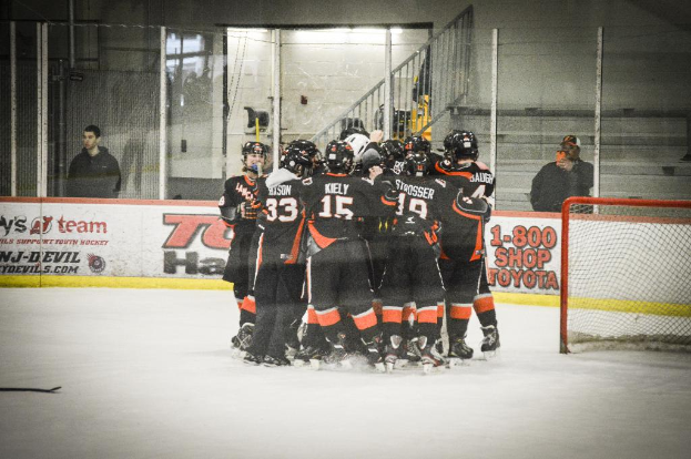 Eine Gruppe von Menschen in Schlittschuhen, Handschuhen und Helmen steht auf einem Eisfeld mit Absperrungen und Glastüren dahinter, ein Hockey-Tor mit Netz auf der rechten Seite und einen Hockey-Schläger auf der linken Seite.