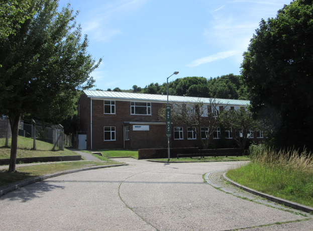 Ein Schulgebäude mit Fenstern, einem Straßenschild, einer Tafel, einer Baumgruppe, Gras, einem Zaun und einem bewölkten Himmel, das inmitten eines Parks umgeben von saftigem Gras liegt.