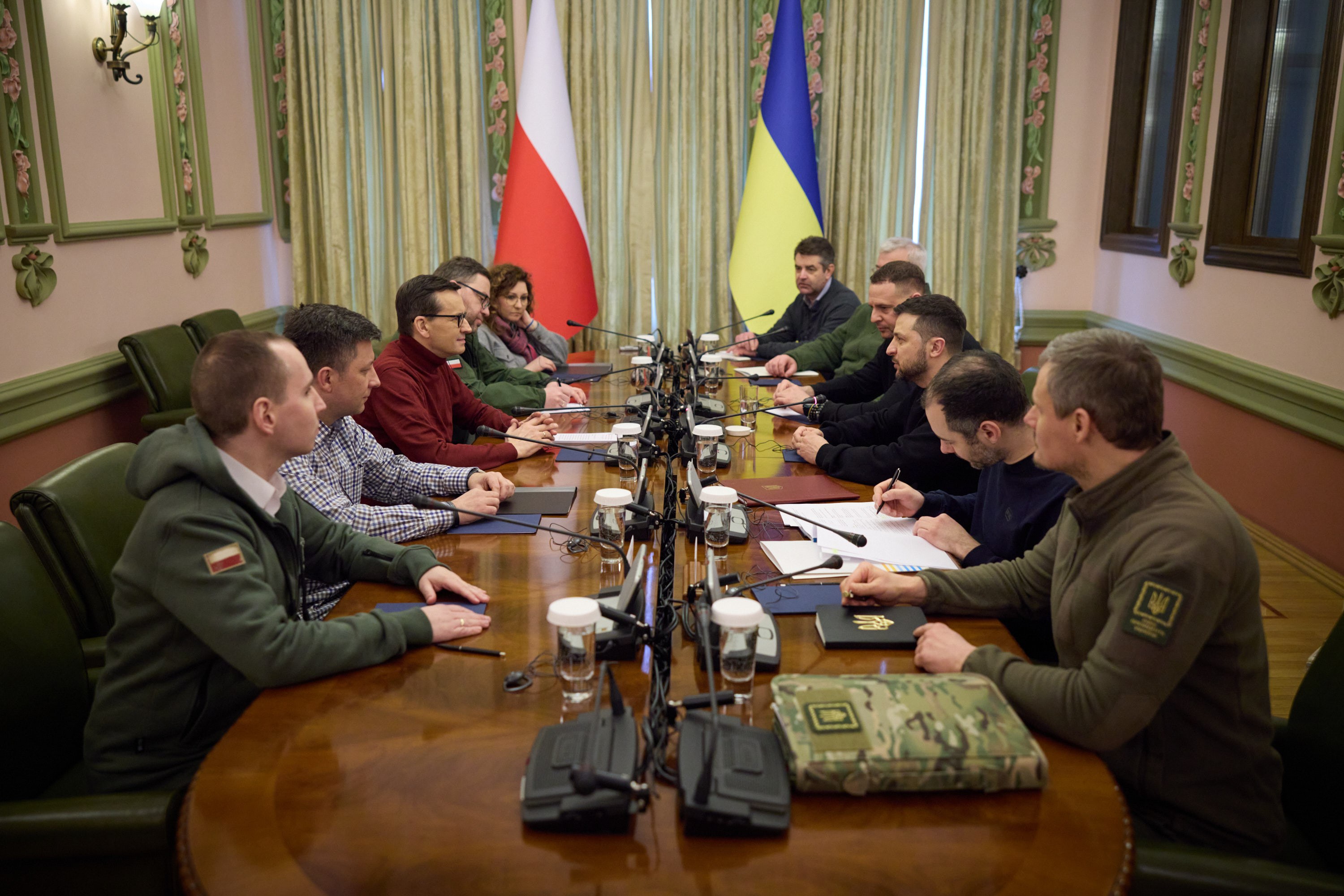 Eine Gruppe von Männern in einer formellen Umgebung mit Mikrofonen, Papieren und Büchern auf einem Holztisch, die intensiv diskutieren mit Flaggen und Lichtern im Hintergrund.