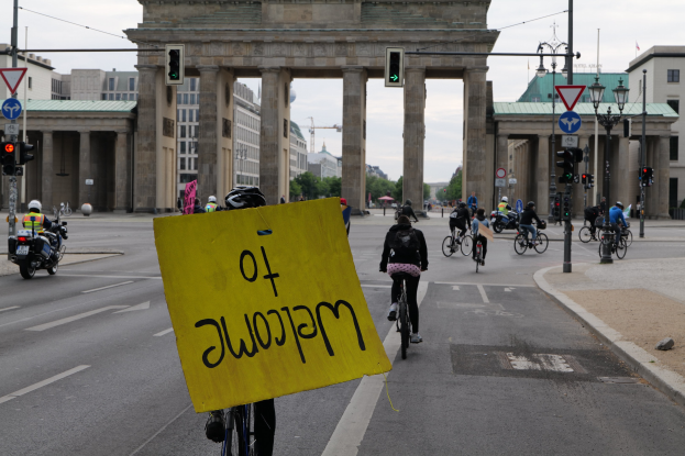 Eine Gruppe von Menschen, die auf Fahrrädern am Brandenburger Tor in Berlin, Deutschland, vorbei fährt, wobei einer ein gelbes Schild hält, unter einem klaren blauen Himmel.