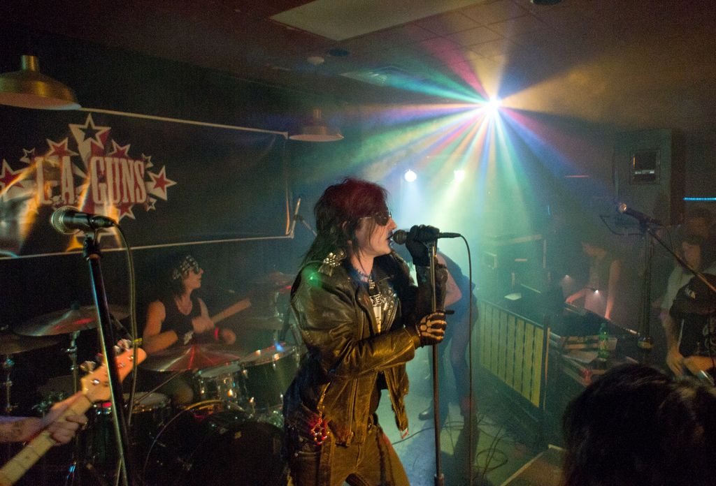 Eine Frau in einer schwarzen Jacke singt in ein Mikrofon auf der Bühne, mit einem Mann in Sonnenbrille und einem Stirnband, der ein Instrument spielt, und einem Plakat mit "LA Guns" im Hintergrund, unter Diskoleuchten und Hängelampen.