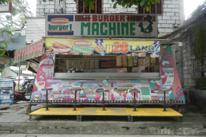 Ein bunt bemalter gelber und roter Foodtruck in Form eines Jumbo-Burgers, der auf einer von Gebäuden, Bäumen und Menschen gesäumten Straße geparkt ist.