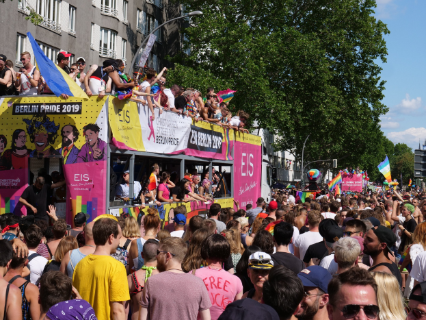 Eine große Menge Menschen, die auf der Straße bei der Berlin Pride 2019 gehen, viele tragen Kappen und Schutzbrillen, einige halten Fahnen, mit Bannern, Laternenmästen, Bäumen und Gebäuden im Hintergrund bei einem bewölktem Himmel.