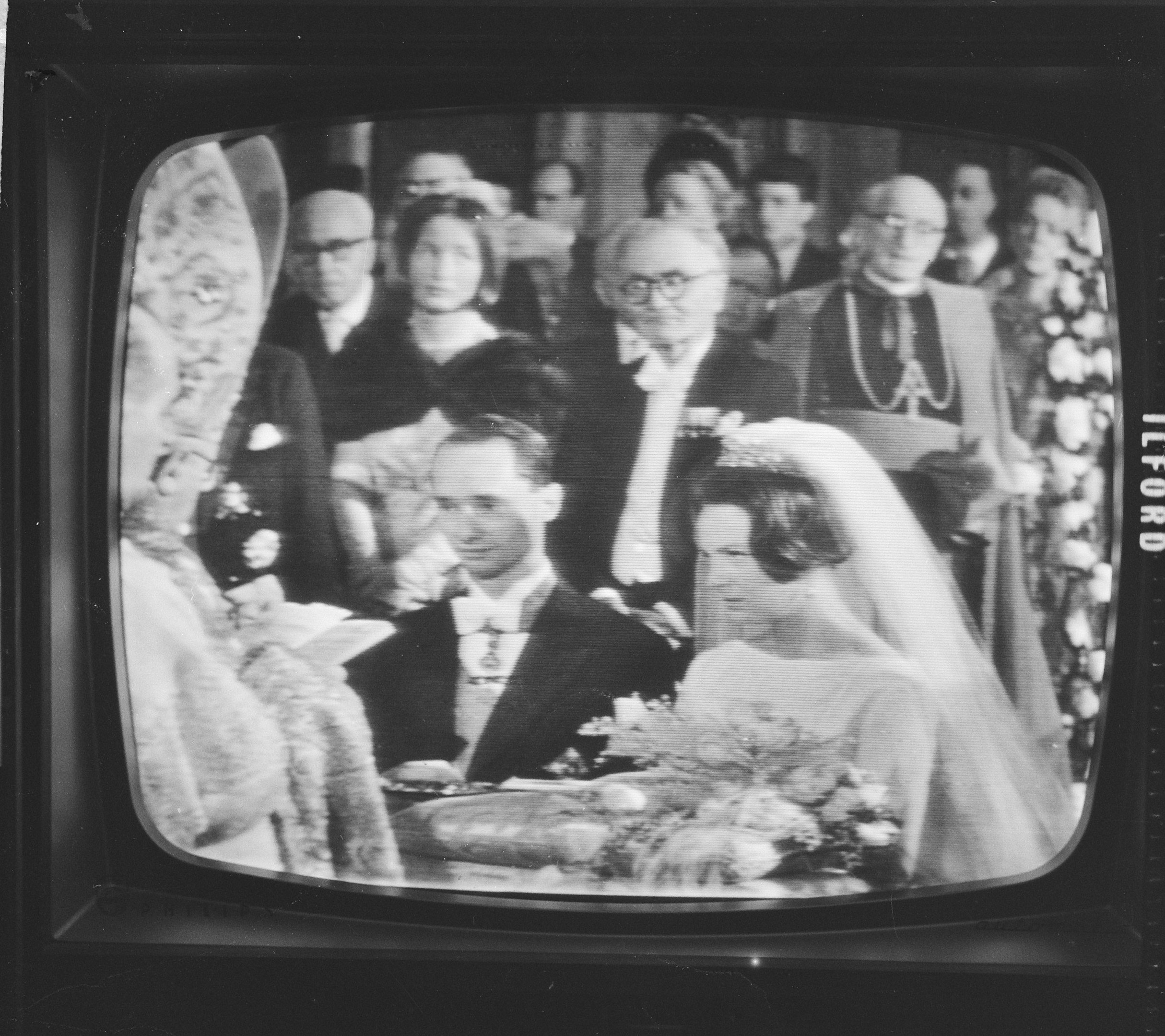 Schwarzes und weißes Foto einer Braut in einem weißen Kleid und Schleier, die einen Strauß hält, neben einem Bräutigam in Anzug und Krawatte steht, auf einem Fernsehbildschirm umgeben von Menschen.