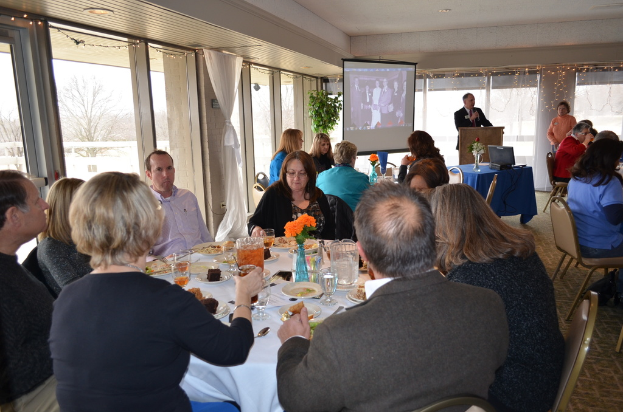 Eine Gruppe von Menschen, die um einen Tisch sitzen, der mit verschiedenen Speisen gefüllt ist, eine Person, die steht und spricht, ein Projektor, der Inhalt anzeigt, eine Pflanze neben dem Projektor und ein Fenster mit Vorhängen, Himmel und Bäumen im Hintergrund.