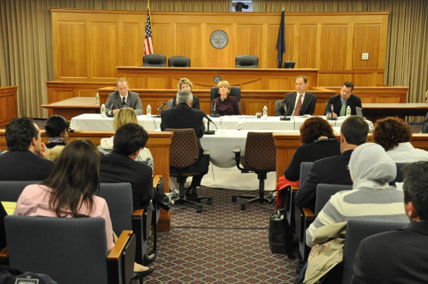 Ein Versammlungsraum mit einer Gruppe von Menschen, die auf Stühlen sitzen, zwei ausländischen Flaggen, einem orangefarbenen Schreibtisch mit Wasserflaschen und einem Mikrofon.