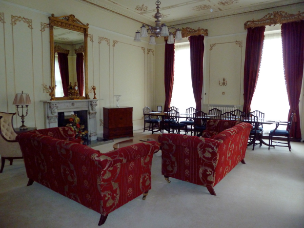 Zwei rote Sofas in einem Wohnzimmer, ein kleiner Teetisch mit einem Buch darauf, ein wandmontierter Spiegel, eine Lampe auf einem Tisch, ein Blumenstrauß vor einem Kamin, ein Esstisch mit Stühlen, von der Decke hängende Lampen, Vorhänge an einer Stange und ein leerer Stuhl.