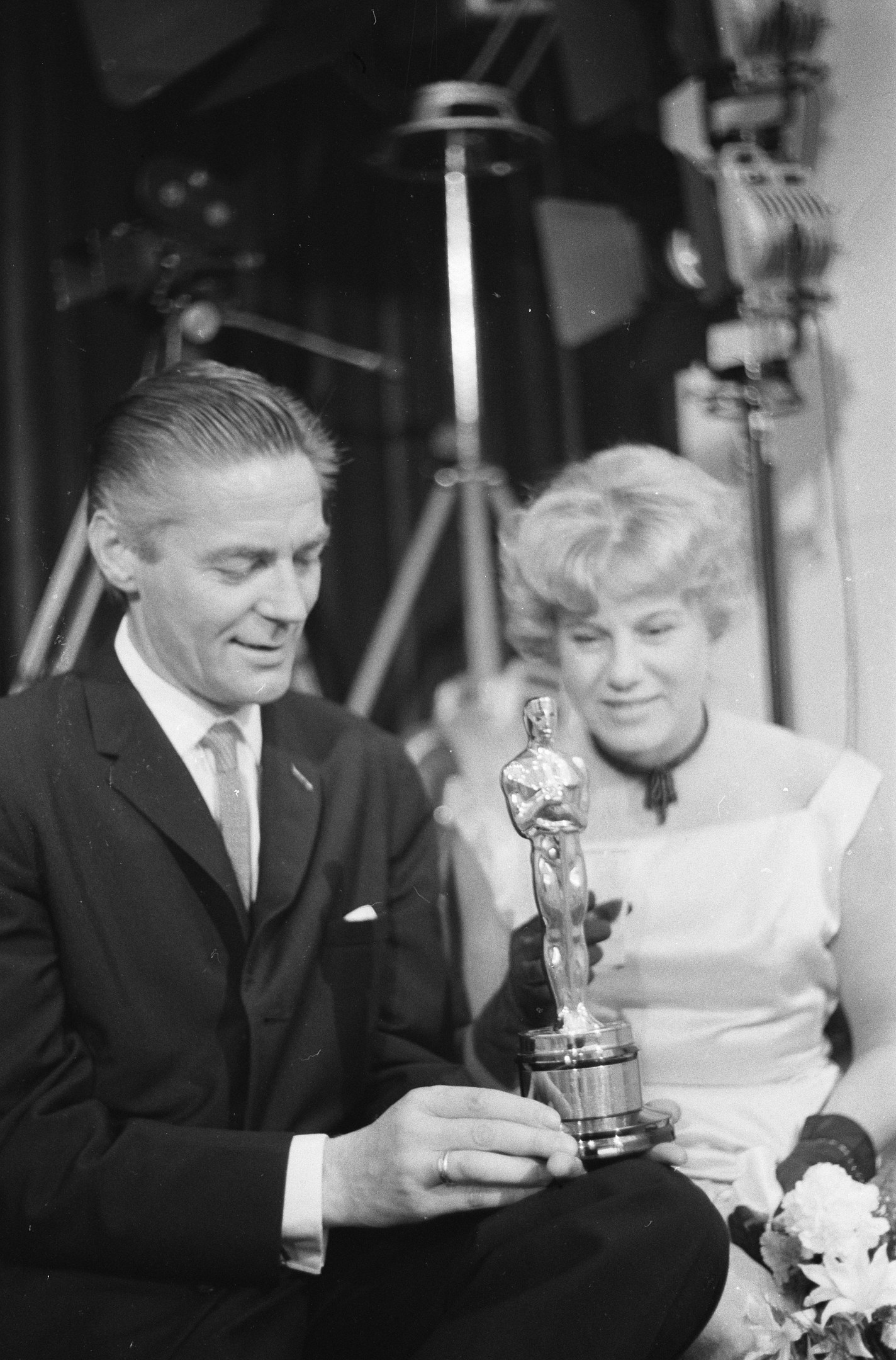 Schwarzes und weißes Foto eines Mannes in einem schwarzen Blazer, weißem Hemd und Krawatte, der eine Oscar-Statue hölt, und einer Frau in Abendgarderobe, die einen Blumenstraußl hölt, nebeneinander sitzend, mit Hintergrundbeleuchtung.