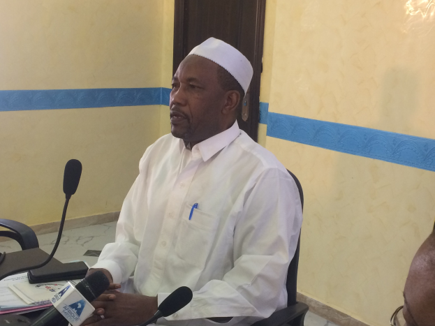 Ein Mann in einem weißen Kleid und Hut sitzt an einem Tisch mit Mikrofonen, spricht, während Papiere und Gegenstände auf dem Tisch liegen, mit einem anderen Mann mit Brille zu seiner Rechten, in einem Raum mit einer Wand und einer Tür im Hintergrund.