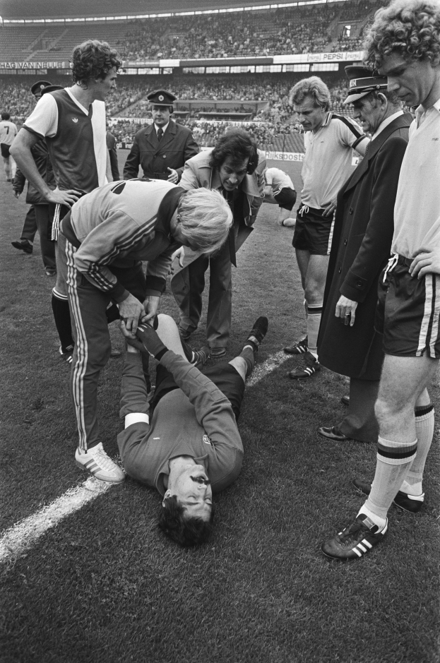 Schwarzes und weißes Bild eines Fußballspielers auf dem Boden umgeben von einer Gruppe von Männern, mit einer Stadionmenge im Hintergrund; der Spieler scheint verletzt zu sein.