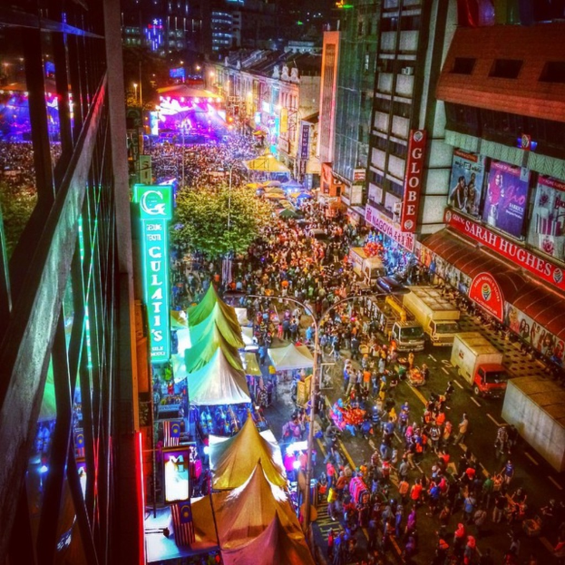 Eine belebte Karnevalsszene mit einer großen Menschenmenge auf der Straße, umgeben von Zelten, Geschäften, Gebäuden und Bannern, mit hell erleuchteten Bereichen im Hintergrund.