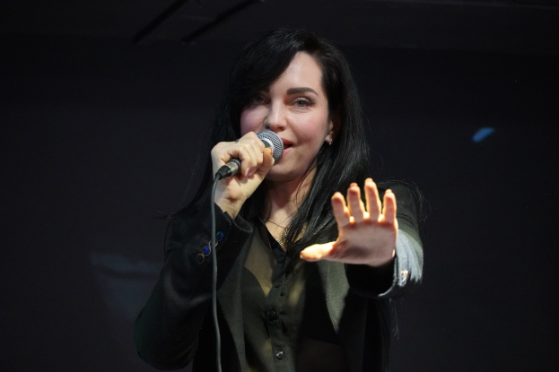 Eine Frau in einem schwarzen Kleid singt in ein Mikrofon auf einer B├╝hne, ihre Augen geschlossen und ihr Gesichtsausdruck voller Emotion und Konzentration, beleuchtet von einem hellen Licht.