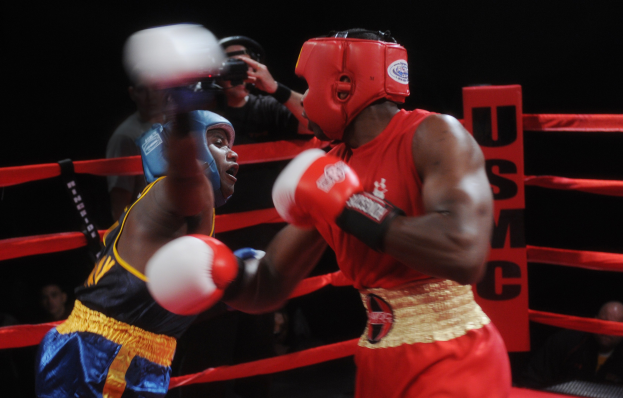 Zwei Boxer in einem Ring, die in einem Kampf gegeneinander antreten, mit einem Fotografen und Zuschauern im Hintergrund bei dramatischer Beleuchtung.