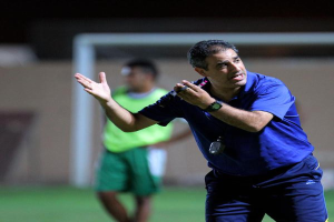 Ein Mann in einem blauen T-Shirt und Hose steht auf einem Fußballfeld und hält ein Mikrofon in der Hand, während er zu seinen Spielern gestikuliert. Im Hintergrund ist eine Person in einem grünen Kleid, ein Torpfosten und eine Wand zu sehen.