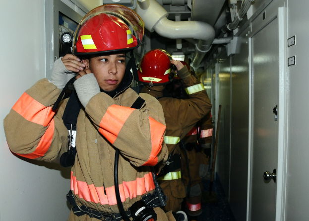 Gruppe von Feuerwehrleuten in Uniform, die in einem Übungsraum mit Rohren und Equipment im Hintergrund stehen.