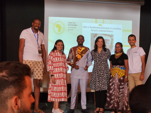 Eine Gruppe von Menschen auf einer Bühne, die für ein Foto posieren und lächeln, mit einem Publikum davor und einem Bildschirm im Hintergrund, der 'Open the Knowledge Journalism Awards 2019' anzeigt.