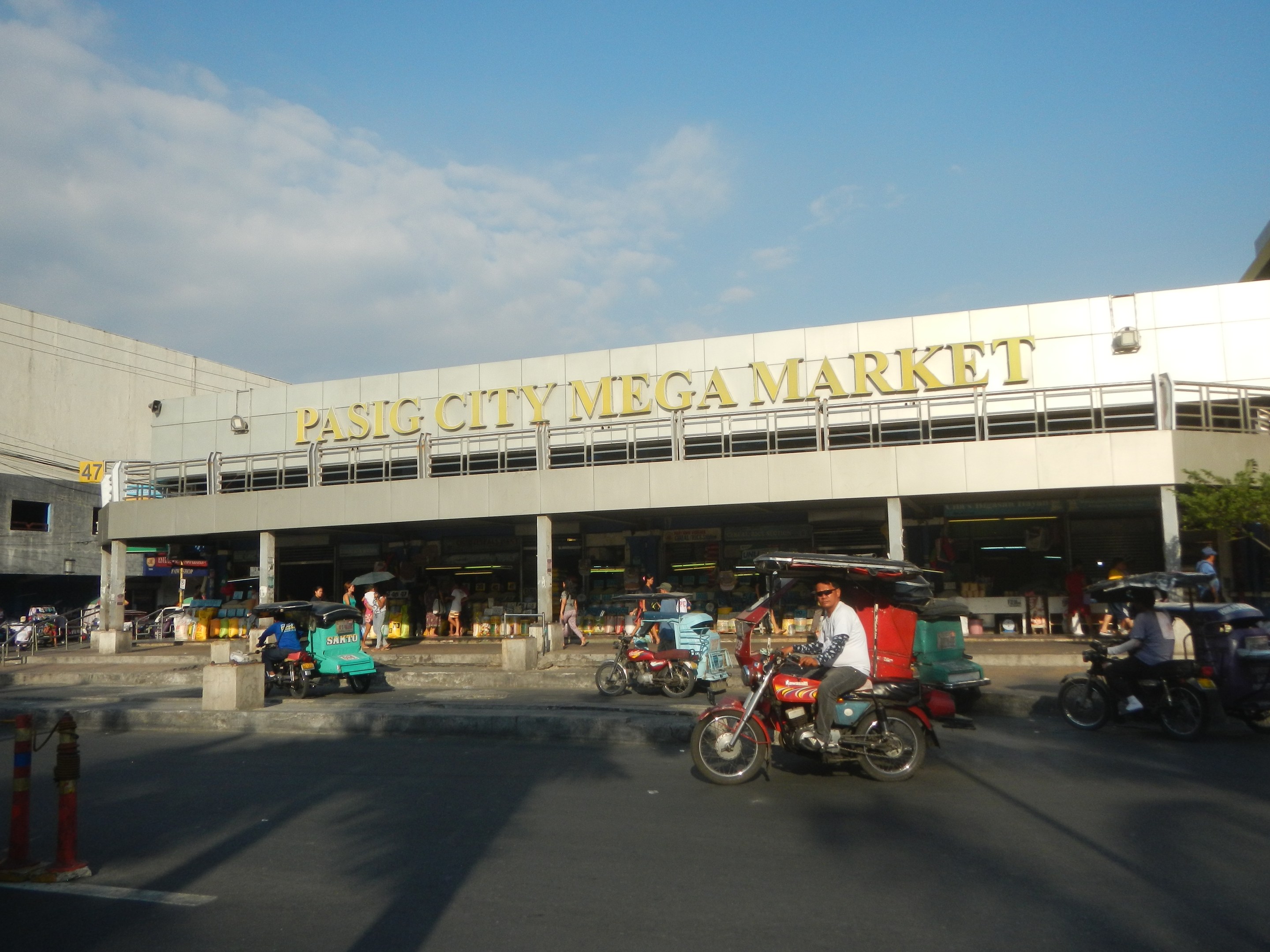 Eine belebte Straßenszene auf dem Pasig City Mega Market mit Fahrzeugen und Fußgängern, ein Gebäude mit Text im Hintergrund und ein bewölkter Himmel.