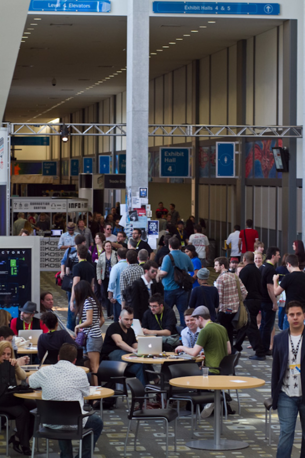 Ein belebter Flughafenvorplatz mit wartenden Menschen, einige stehen und andere arbeiten, Richtungsschilder an der rechten Wand und √∂ber ihnen sowie von der Decke h√§ngende Lichter.