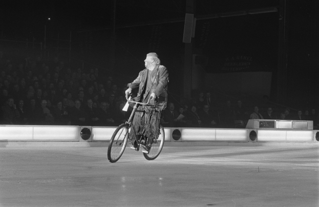 Schwarz-weißes Bild eines Mannes, der auf einem Eisstadion Fahrrad fährt, im Hintergrund sitzen einige Menschen und eine Wand ist zu sehen.