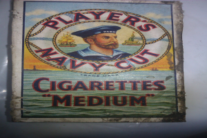 Ein Vintage-Plakat zeigt eine ernste Person in einer marineblauen Uniform mit weißem Hemd und roter Krawatte, die Arme verschränkt, mit der Aufschrift "Player's Navy Cut Cigarettes Medium" in fetter schwarzer Schrift auf einem weißen Hintergrund.