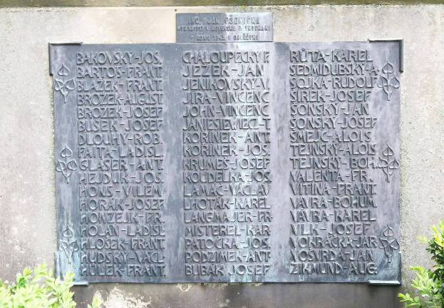 Ein Steintafel an einer Wand mit eingraviertem Text, umgeben von Pflanzen unten, wahrscheinlich ein Holocaust-Mahnmal.