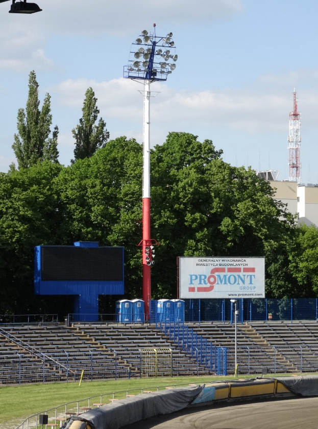 Rennstrecke mit Überdachung, umgeben von Gras, Geländern, Stufen, einem Lichtmast, einem Schild, Bäumen, Gebäuden, Türmen und einem bewölkten Himmel.
