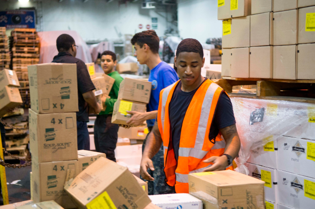 Eine Gruppe von Männern in orangefarbenen Westen und Sicherheitswesten lädt Kisten in ein Lagerhaus, mit mehreren Kartons auf der rechten Seite und einer Kette auf der linken Seite und einer beleuchteten Wand im Hintergrund.
