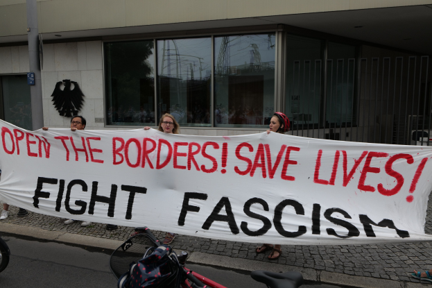 Gruppe von Menschen mit einem Banner "Öffnet die Grenzen, rettet Leben, kämpft gegen Faschismus" vor einem Gebäude mit Glasfenstern und einer Stange, mit einem Fahrrad und einer Tasche auf der Straße.
