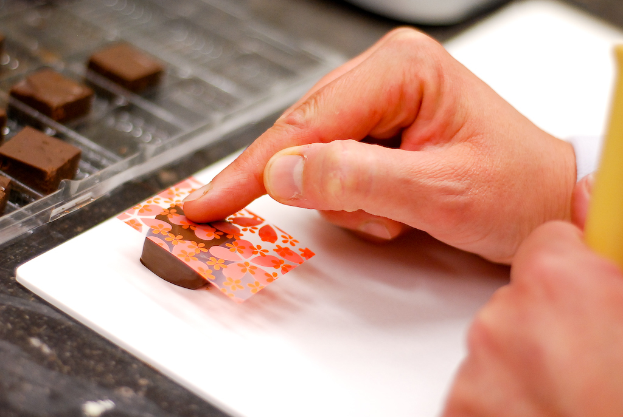 Eine Hand, die eine dekorative Abdeckung auf eine Schokolade legt, mit weiteren Schokoladen auf einem Tablett links und einem Tisch darunter.