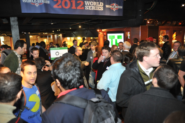 Eine große Menschenmenge bei einem Event mit einem Bildschirm, der ein Bild anzeigt, sowie Lichtern und einer Holzwand im Hintergrund.