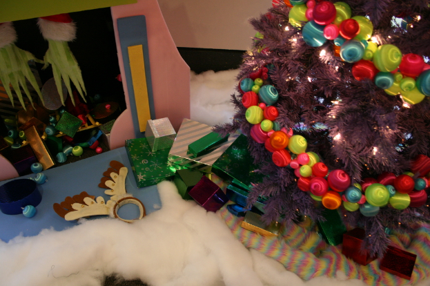 Eine Indoor-Szene mit einer Wand im Hintergrund, einem Baumwoll- und Wolltuch unten, einem geschmückten Weihnachtsbaum rechts, Geschenkboxen und Dekorationsgegenständen in der Mitte und einer Hütte mit Dekorationsgegenständen und Baumwolle oben links.