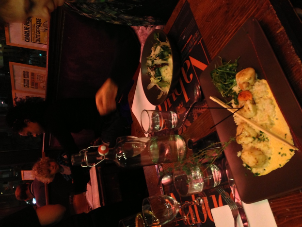 Ein Tisch mit verschiedenen Speisen, Tellern, Besteck und Papieren, mit Menschen und Brettern im Hintergrund.