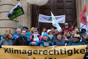Eine Gruppe von Menschen steht vor einem Gebäude mit Säulen und einer Tür, hält ein Banner mit Text, Fahnen mit Stangen und Ballons und nimmt an einem Klimastreik in Deutschland teil.